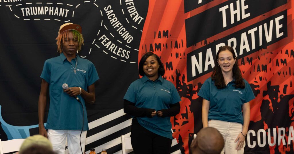Three community tech workers stand on stage holding a microphone with banner behind them for Digital Inclusion Week