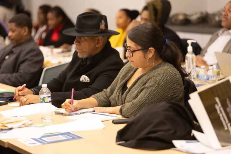 Latina entrepreneur takes notes during a workshop sititng next to a black man in a fedora
