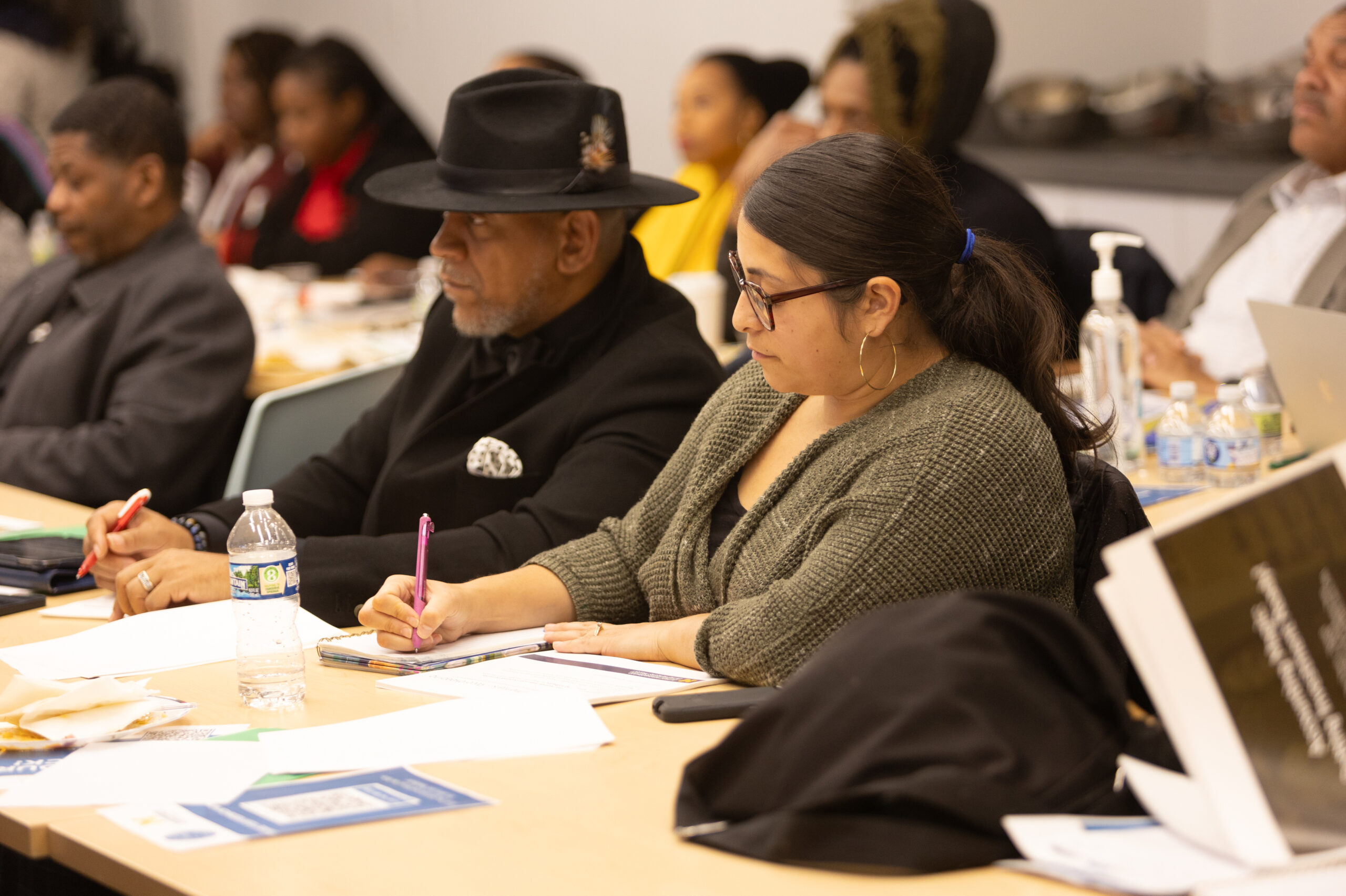 Latina entrepreneur takes notes during a workshop sititng next to a black man in a fedora