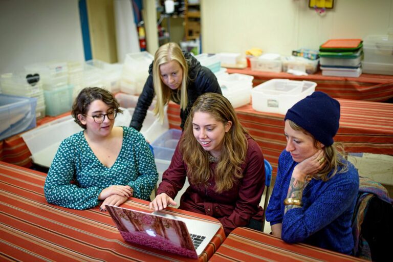 three white women look at a laptop while a fourth leans over behind them they are looking focused