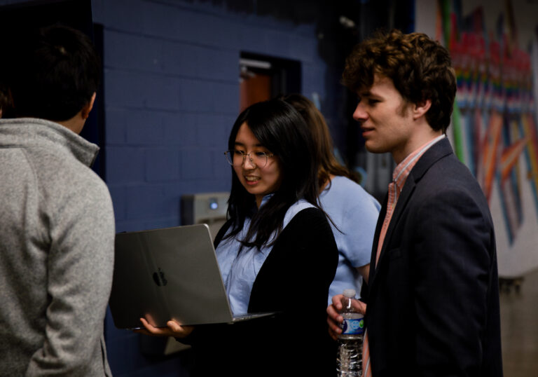 asian woman holds a laptop and smiles while white man looks on dark blue walls in background