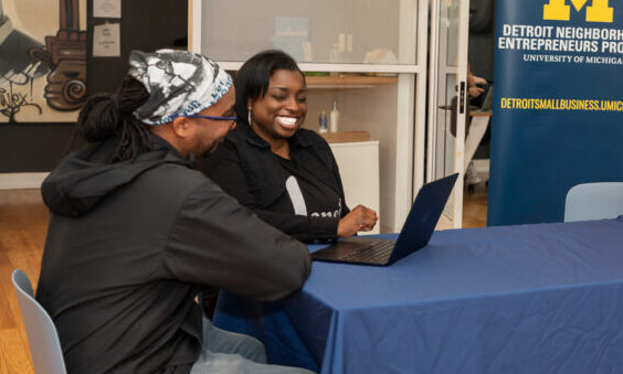 black man sits at a table looking on while black woman entepreneur smiles wide with her hand over laptop